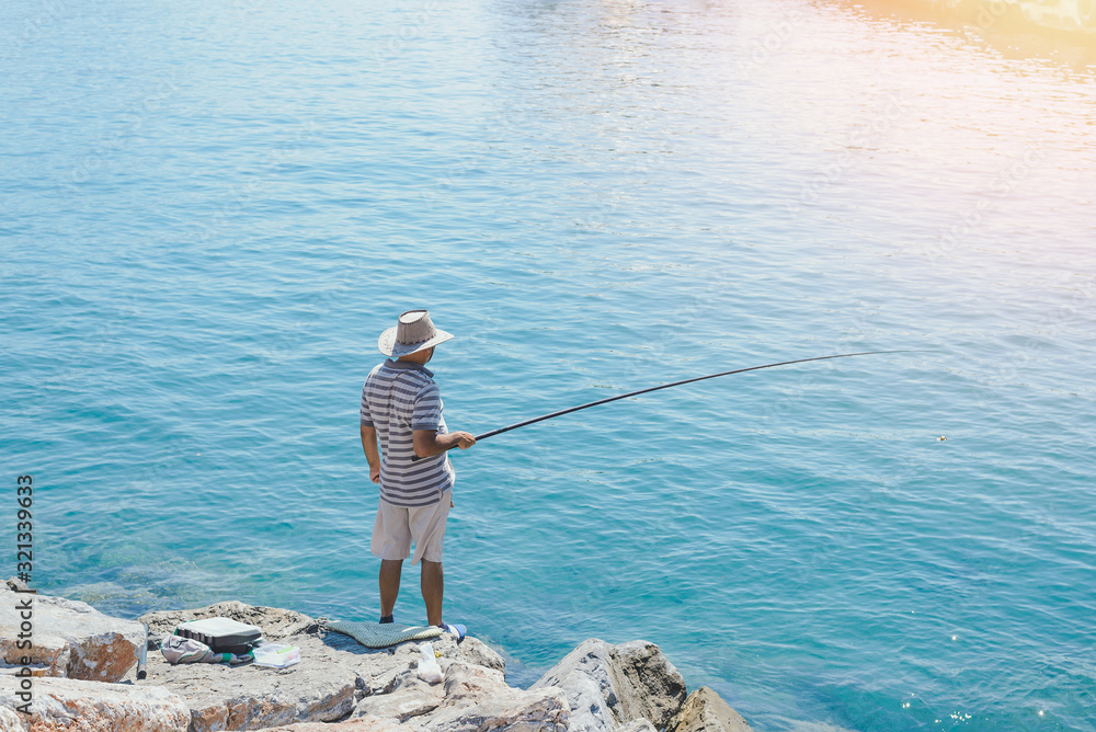 Fisherman in big hat is fishing on sea rocks alone in sunny day.