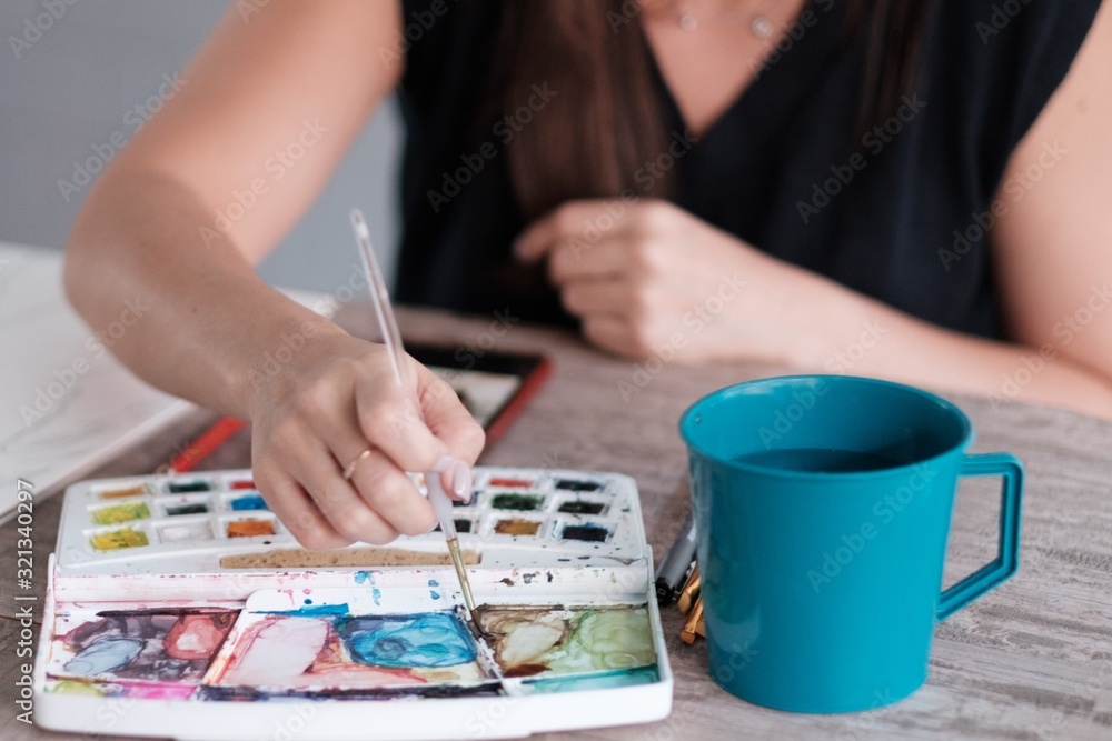 Fototapeta premium Unrecognizable woman mixing watercolors with her thin brush. There's a cup of water next to her paint that she can use to clean her brush.