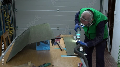 specialist worker with protections on face and hands cutting piece of carbon fiber - kevlar with cutting machine and vacuuming the resulting powder at workshop. Manufacturing part for racing car