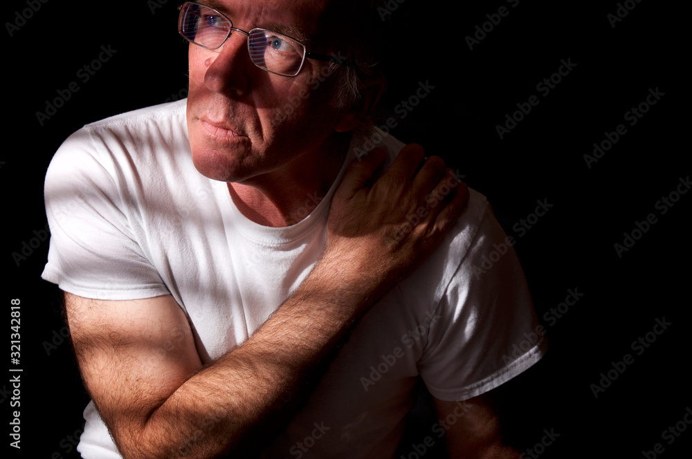 Blue eyed man looking away, holding shoulder and sitting in a dark room.