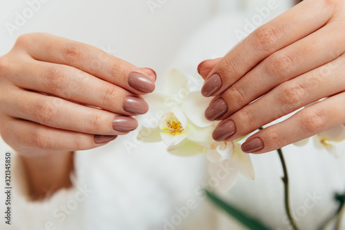 Beige manicure gel polish on beautiful oval nails of a woman in a white sweater with an orchid flower.