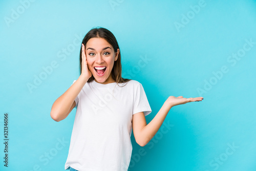 Young caucasian woman  isolated holds copy space on a palm, keep hand over cheek. Amazed and delighted.