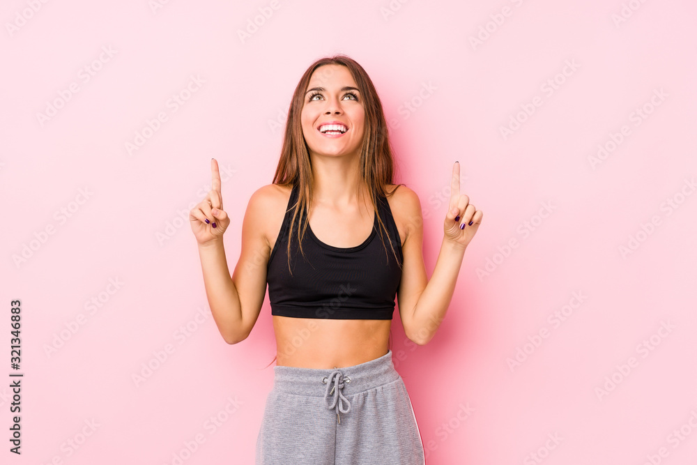 Young caucasian fitness woman posing in a pink background indicates with both fore fingers up showing a blank space. Young caucasian fitness woman posing in a pink background indicates with both fore fingers up showing a blank space.