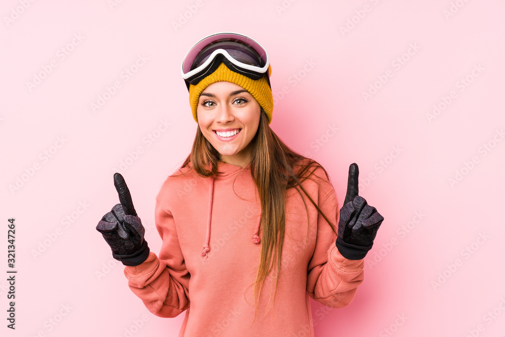 Young caucasian woman wearing a ski clothes isolated indicates with both fore fingers up showing a blank space. Young caucasian woman wearing a ski clothes isolated indicates with both fore fingers up showing a blank space.