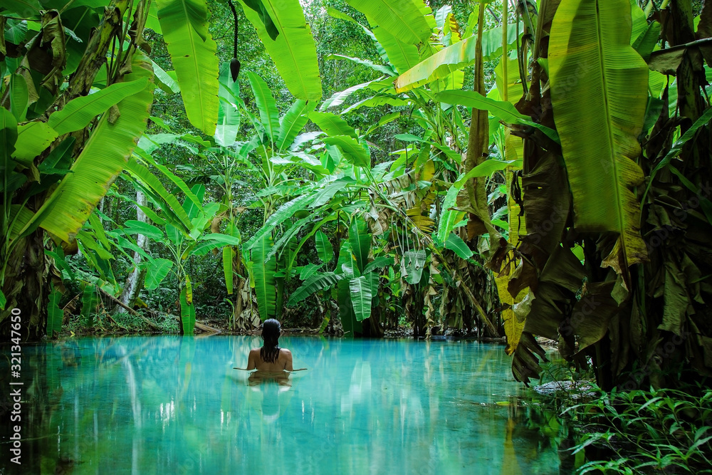 A girl stands in a natural pool surrounded by banana tree's leafs - obrazy, fototapety, plakaty 