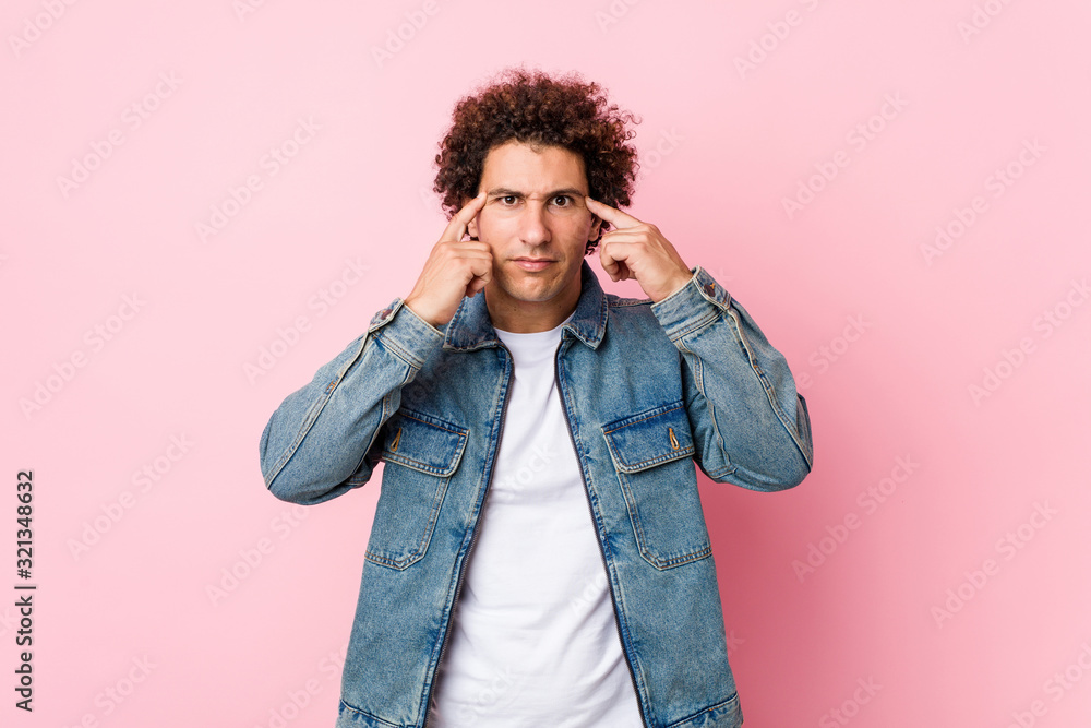 Curly mature man wearing a denim jacket against pink background focused on a task, keeping forefingers pointing head. Curly mature man wearing a denim jacket against pink background focused on a task, keeping forefingers pointing head.