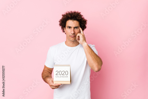 Papier peint Young caucasian curly man holding a 2020 calendar pointing his temple with finger, thinking, focused on a task