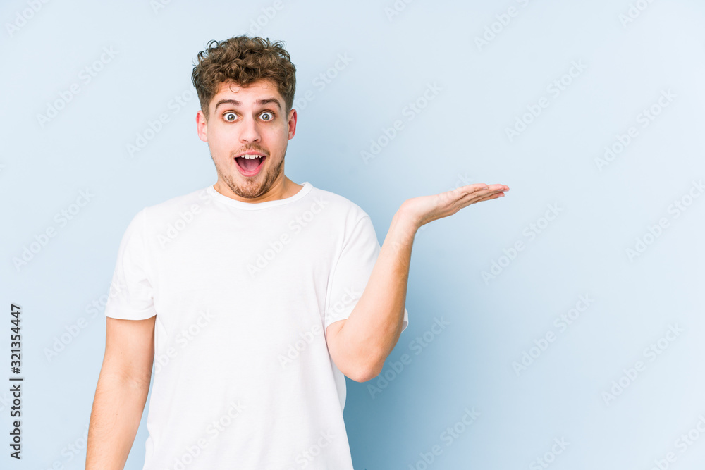 Young blond curly hair caucasian man isolated impressed holding copy space on palm. Young blond curly hair caucasian man isolated impressed holding copy space on palm.