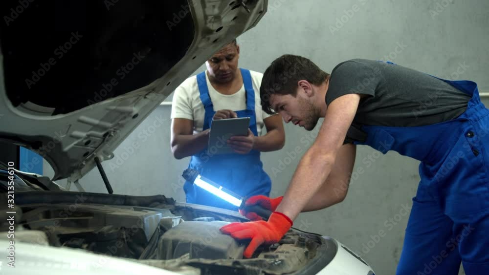 Mechanic with flashlight examines the motor of car and his collegue takes notes