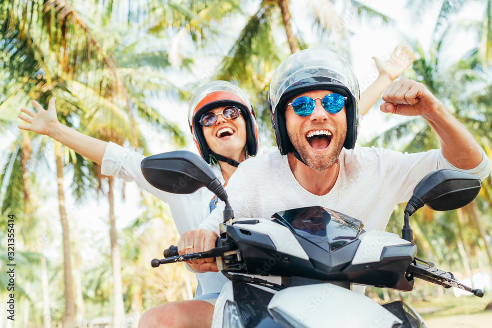 Happy smiling couple travelers riding motorbike scooter in safety helmets during tropical vacation under palm trees on Ko Samui island in Thailand - obrazy, fototapety, plakaty 