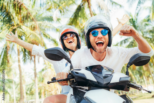 Happy smiling couple travelers riding motorbike scooter in safety helmets during tropical vacation under palm trees on Ko Samui island in Thailand