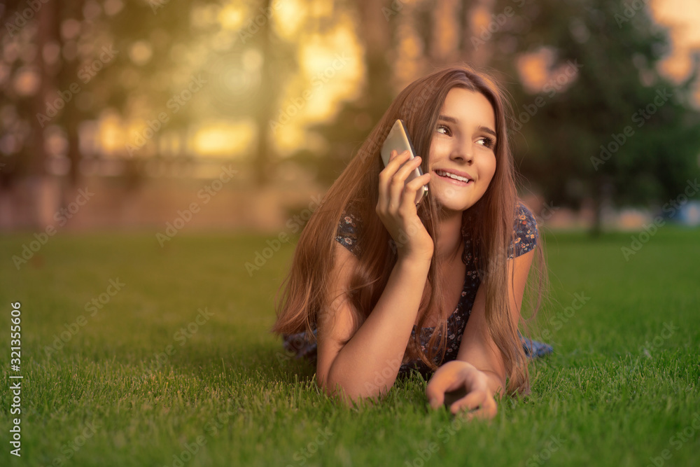 Happy at phone. Smiling young woman talking at cell mobile phone and lying on meadow grass lawn in the city trees on background in the summer evening. Happy at phone. Smiling young woman talking at cell mobile phone and lying on meadow grass lawn in the city trees on background in the summer evening.