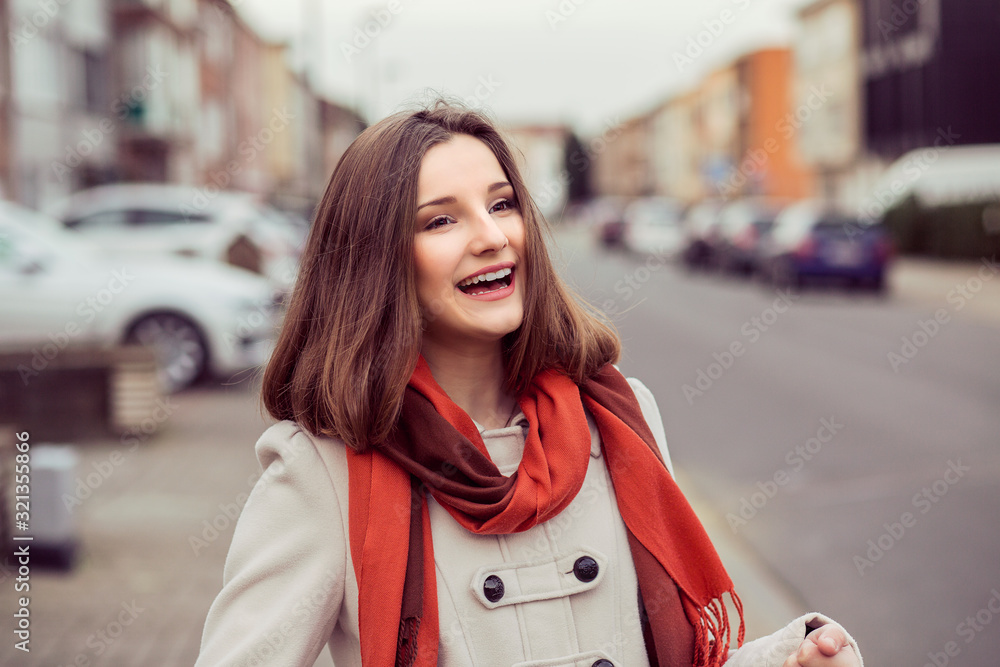 Smiling woman out at evening in the city looking away from camera, portrait in autumn spring wearing beige coat, red scarf