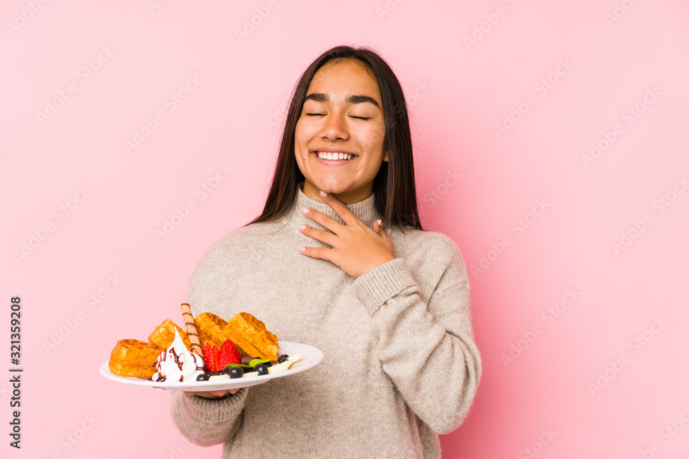 Young asian woman holding a waffle isolated laughs out loudly keeping hand on chest.