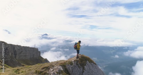 Aerial view. Young man hiking at Seceda mountain raising arms to the sky on the peak at sunrise. Backpack, yellow jacket, boots, beanie. Traveling to puez Odle, Dolomites, Trentino, Italy.