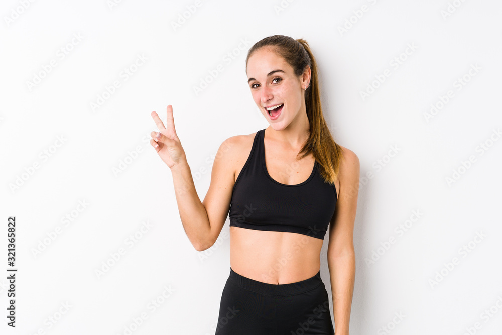 Young caucasian fitness woman posing in a white background joyful and carefree showing a peace symbol with fingers. Young caucasian fitness woman posing in a white background joyful and carefree showing a peace symbol with fingers.