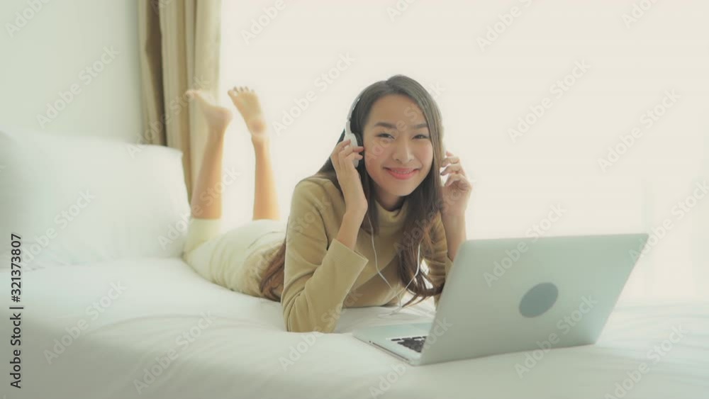 A young woman lying prone on a bed sways to the music she is listening to through headphones attached to her laptop. A young woman lying prone on a bed sways to the music she is listening to through headphones attached to her laptop.