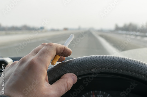close up of a driver's hand holding a cigarette and steering wheel of a car while driving on a highway, country road