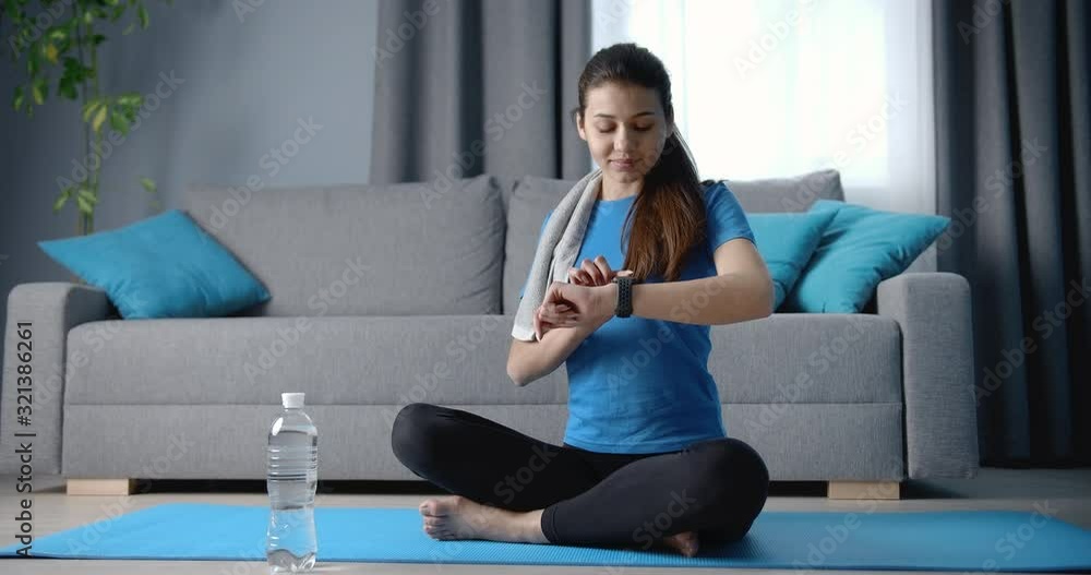 Cheerful young lady with long dark hair tied in knot using her smart watch for detecting training time at home. Fitness woman in sport clothing relaxing on yoga mat with crossed legs. Cheerful young lady with long dark hair tied in knot using her smart watch for detecting training time at home. Fitness woman in sport clothing relaxing on yoga mat with crossed legs.