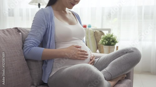 pregnancy and motherhood concept. focus on huge belly of pregnant woman sitting on couch and hands touching in sunlight living room. young future female parent feeling unborn baby in big tummy.