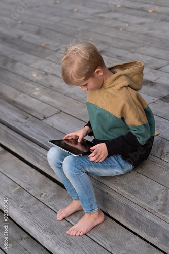 A small blond boy uses a tablet while sitting on a warm pier barefoot