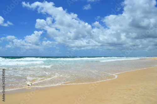 Gentle waves are breaking onto the smooth sand of a beach. A white shell is on the tide line. The sky is blue, with white clouds. Small figures are in the distance.