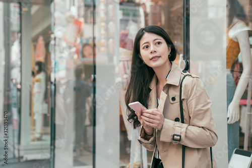 Young asian japanese woman using smartphone while standing in shopping district of city with a fashion store window in background. smiling girl searching direction on mobile phone in modern urban.