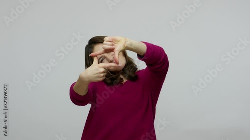 Cheerful brunette woman in pullover looking through photo frame made of fingers, viewing at camera with interest, having fun, pretending to take picture. indoor studio shot isolated on gray background