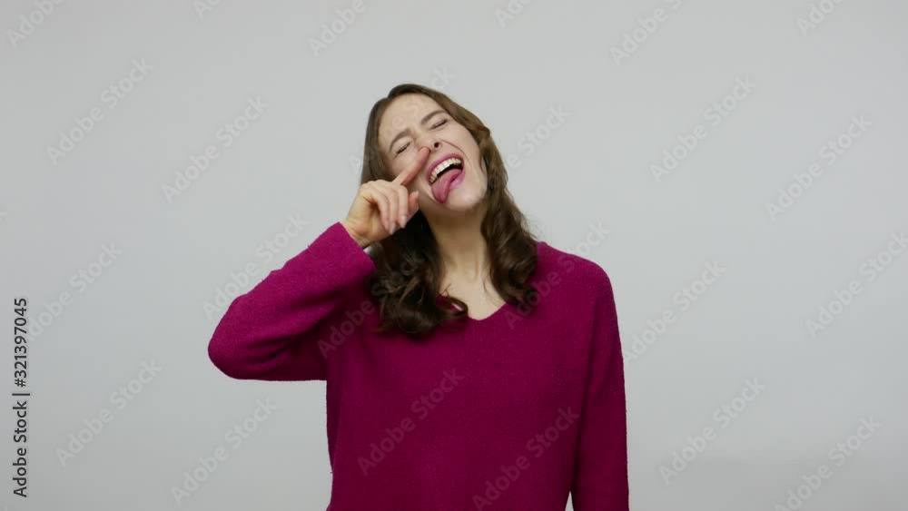Amusing funny brunette woman in pullover laughing and picking her nose with comical foolish humorous expression, sticking out tongue making faces. indoor studio shot isolated on gray background Amusing funny brunette woman in pullover laughing and picking her nose with comical foolish humorous expression, sticking out tongue making faces. indoor studio shot isolated on gray background