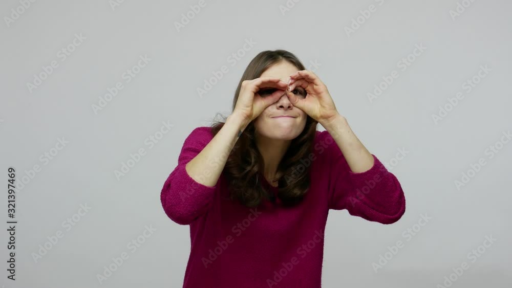 Brunette woman with funny grimace exploring world around, looking through fingers in binoculars gesture with attentive curious expression, smiling when peeking into camera. studio shot gray background Brunette woman with funny grimace exploring world around, looking through fingers in binoculars gesture with attentive curious expression, smiling when peeking into camera. studio shot gray background