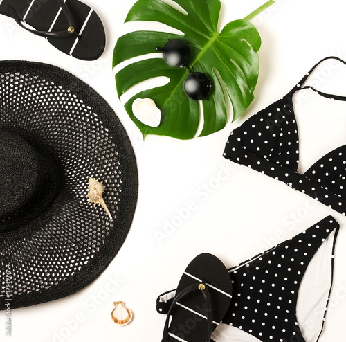 Woman's swimwear and beach accessories flat lay top view on white background. black and white polka dot bikini swimsuit, black straw hat, shoes, sunglasses, monstera leaf, shells. Copy space photo