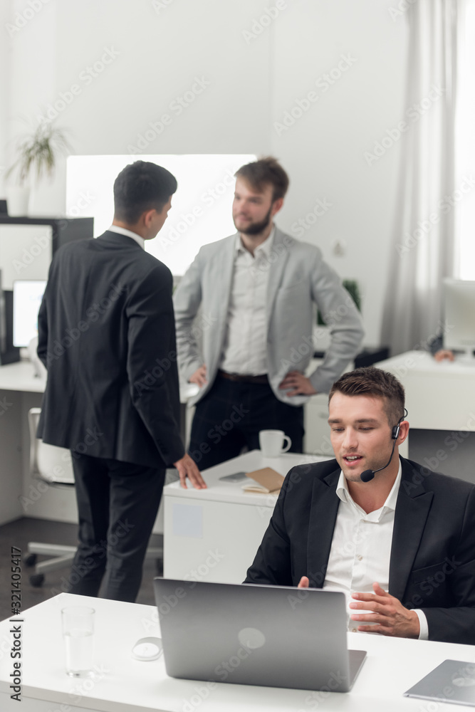 Man with micro have a conversation on laptop, his collegues are talking beside of him Man with micro have a conversation on laptop, his collegues are talking beside of him