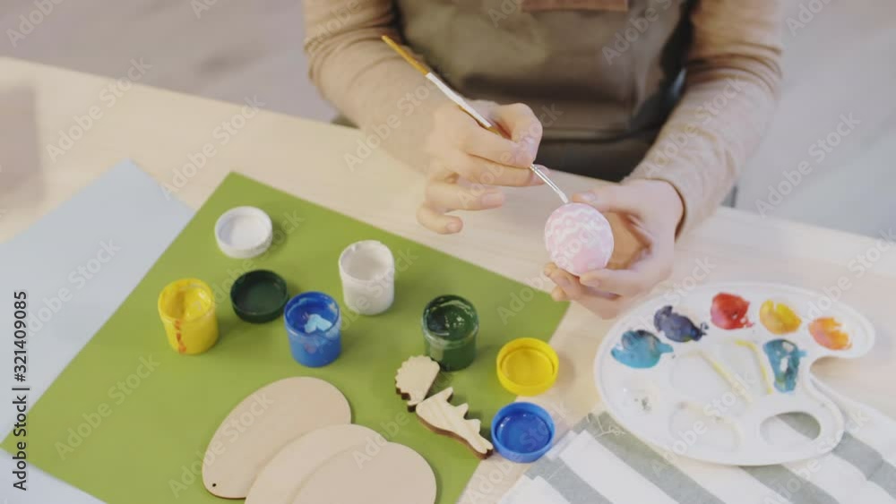 High angle PAN shot of unrecognizable woman using gouache paint and making pattern on pink colored Easter egg. Wooden ornaments and palette lying on table High angle PAN shot of unrecognizable woman using gouache paint and making pattern on pink colored Easter egg. Wooden ornaments and palette lying on table