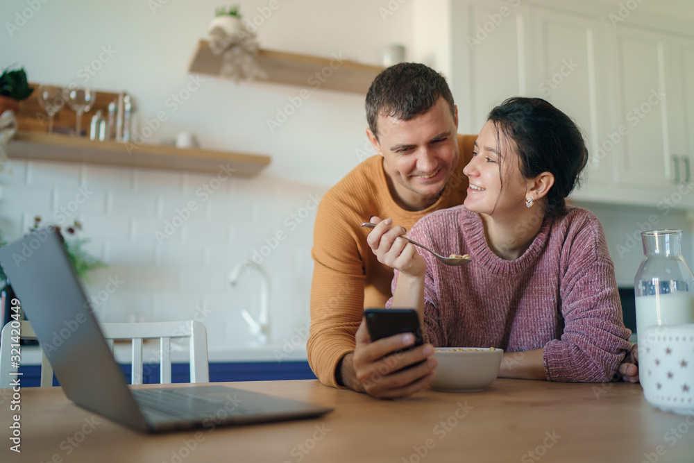 Obraz premium Beautiful young married couple having breakfast together working on laptop at table or reading news. Loving husband and wife making plans for vacation in the kitchen at home