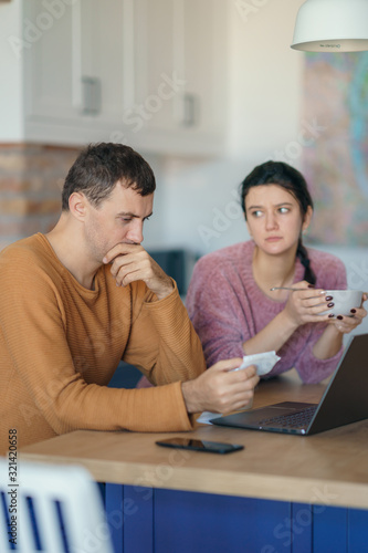Young stressed Caucasian couple facing financials troubles, sitting at kitchen table with bills, checks and laptop computer and reading document from bank, looking frustrated and unhappy