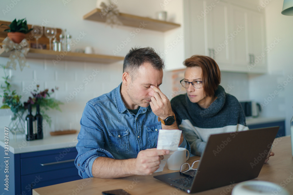 Young stressed Caucasian couple facing financials troubles, sitting at kitchen table with bills, checks and laptop computer and reading document from bank, looking frustrated and unhappy Young stressed Caucasian couple facing financials troubles, sitting at kitchen table with bills, checks and laptop computer and reading document from bank, looking frustrated and unhappy