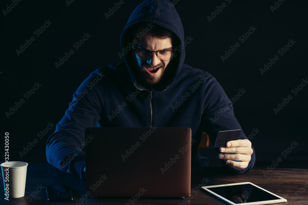 hacker man sit in hood of pullover, male sit excitedly look at screen of laptop after successful hacking, hold bank card in hand, laptop and tablet. man engaged in hacking, commit crime, cyberattack hacker man sit in hood of pullover, male sit excitedly look at screen of laptop after successful hacking, hold bank card in hand, laptop and tablet. man engaged in hacking, commit crime, cyberattack
