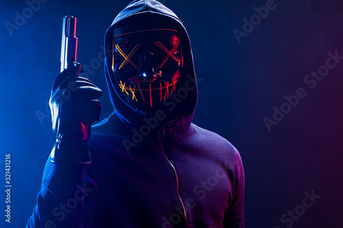 incognito killer in mask hold gun isolated in space with neon lights. dangerous anonymous burglar in the hood of pullover stand and look at camera, wearing scary mask
