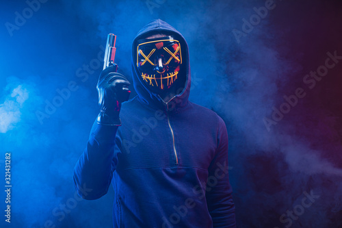 confident dangerous killer male in pullover stand in the hood with gun, anonymous masked hunter man isolated over smoky space with neon lights. crime, kill, hunt, danger concept
