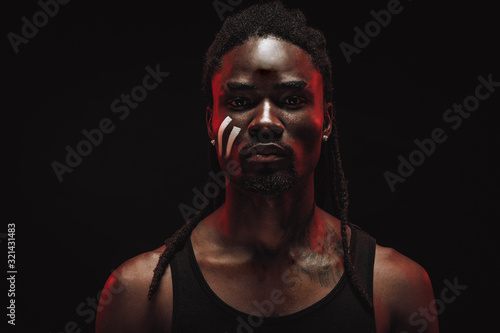 young confident african male in black shirt, sensual bodybuilder seriously look at camera. neon lights falling on his face. black background © Roman