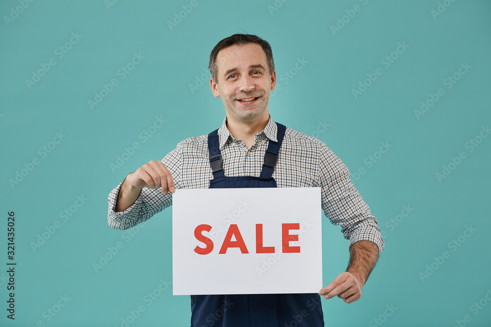Waist up portrait of smiling worker holding SALE sign while posing against blue background, copy space Waist up portrait of smiling worker holding SALE sign while posing against blue background, copy space