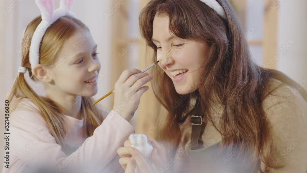 Medium shot of playful preteen girl in bunny ears headband putting paint on nose of her elder sister while decorating Easter ornament Medium shot of playful preteen girl in bunny ears headband putting paint on nose of her elder sister while decorating Easter ornament