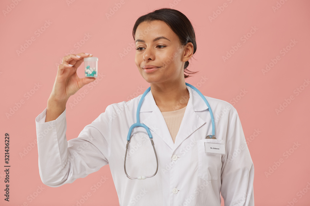 Waist up portrait of beautiful female doctor holding pill bottle and smiling while standing against pale pink background, copy space Waist up portrait of beautiful female doctor holding pill bottle and smiling while standing against pale pink background, copy space