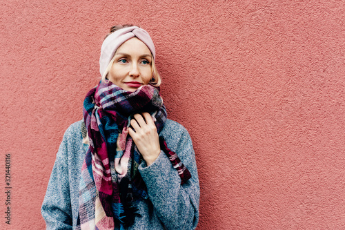 Portrait of a stylish lady in a winter coat with a large plaid scarf and a bandage on her hair against a pink wall.