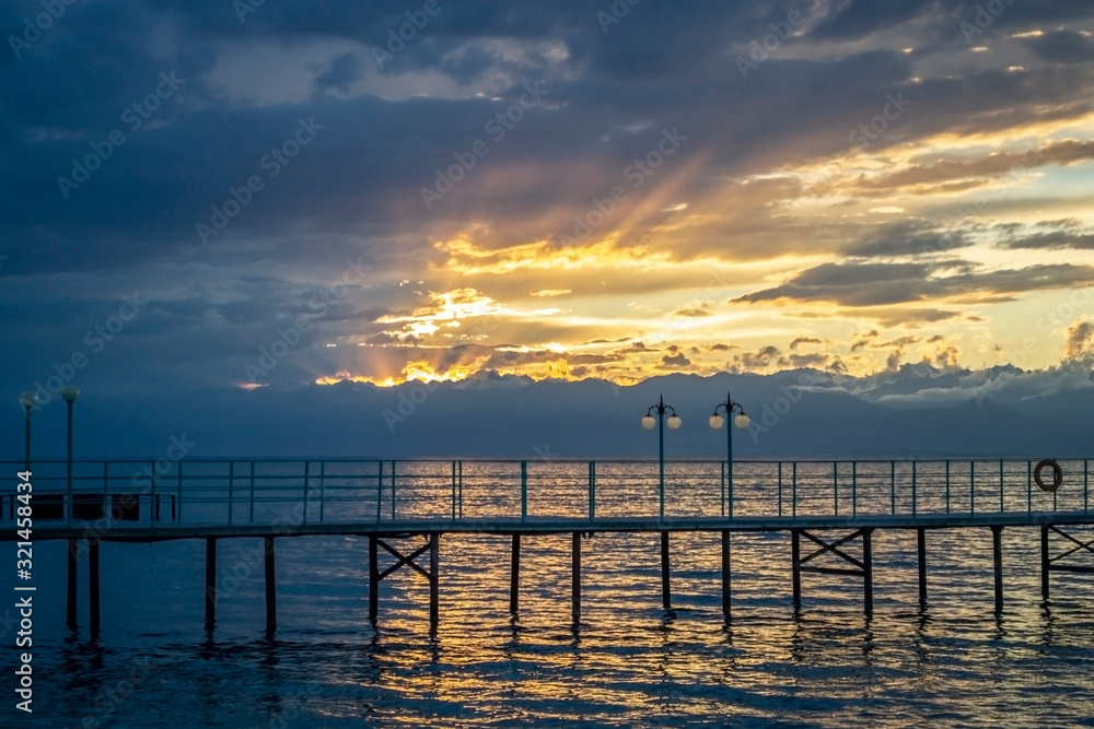 Beautiful sunset on mountain lake with pier, mountains and dramatic cloudy sky with sun rays background.