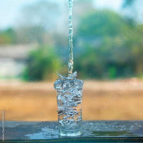 Drink water pouring in to glass over sunlight and natural green background.Water splash in glass Select focus blurred background.Natural Environment concept.