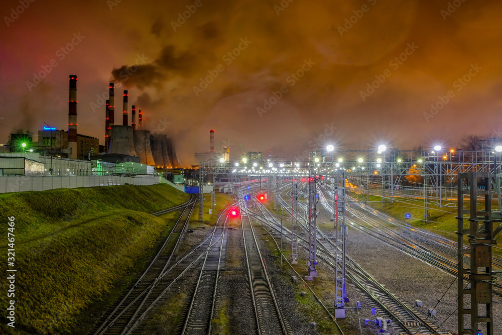 A network of intertwined railroad tracks illuminated by bright illumination at night and pipes of industrial enterprises that spew puffs of smoke into the atmosphere. Industrial landscape showing terr