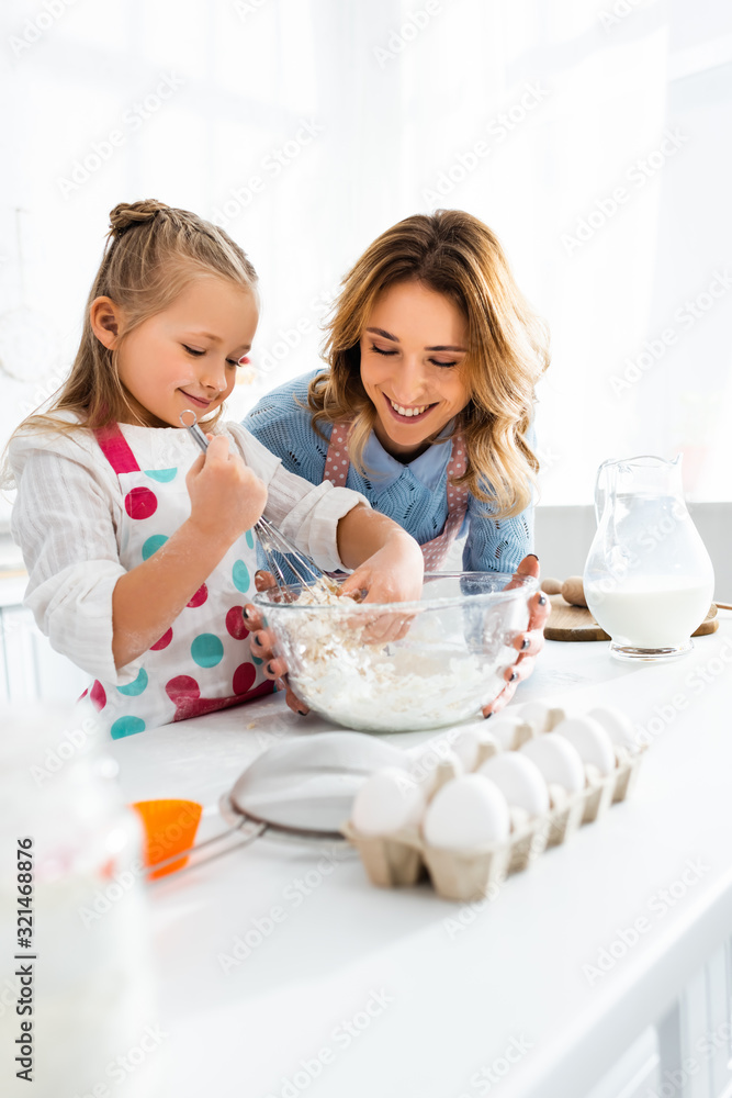 Selective focus of mother looking at daughter kneading dough in bowl at jug of milk, eggs, flour and cooking utensils Selective focus of mother looking at daughter kneading dough in bowl at jug of milk, eggs, flour and cooking utensils