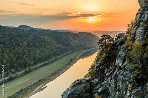 View of the Elbe valley in the Saxon Switzerland National Park. River Elbe at sunset. Colorful sky with yellow-red sun. Landscape with rocks, trees and forests in autumn mood