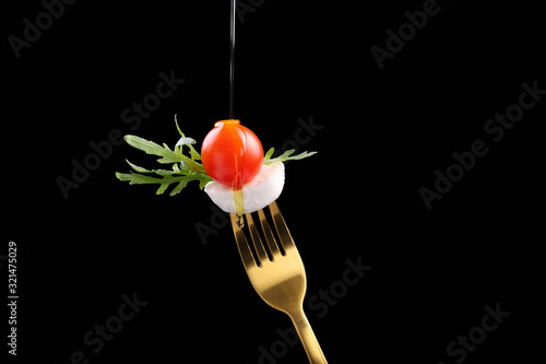 Slice of mozzarella cheese with arugula salad and cherry tomato on a golden fork, watered with olive oil, on a black background.